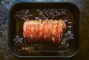 A tied roast pork with crushed grapes in a roasting pan.