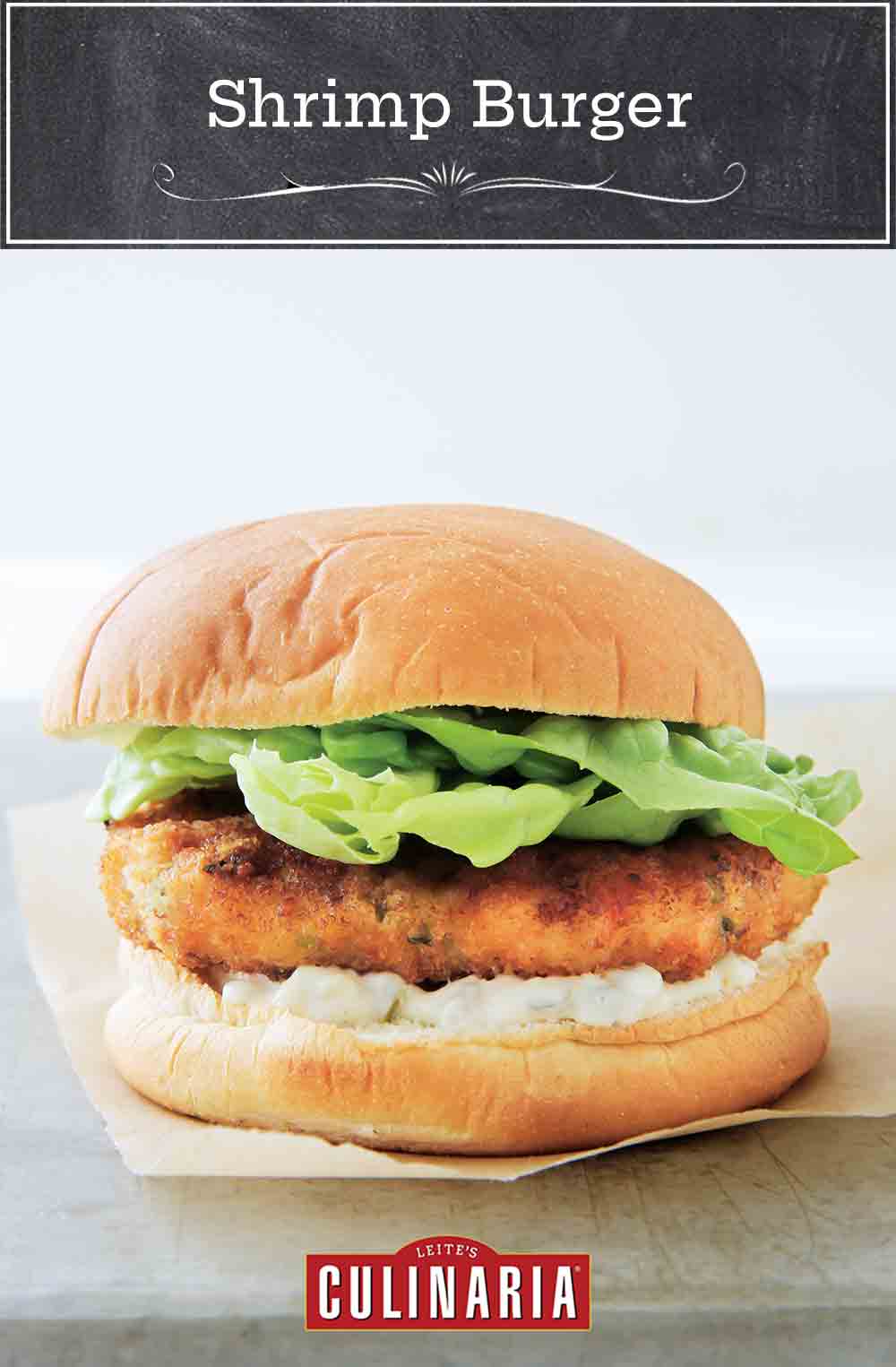 A shrimp burger with tartar sauce, topped with lettuce in a white bun on a piece of parchment paper.