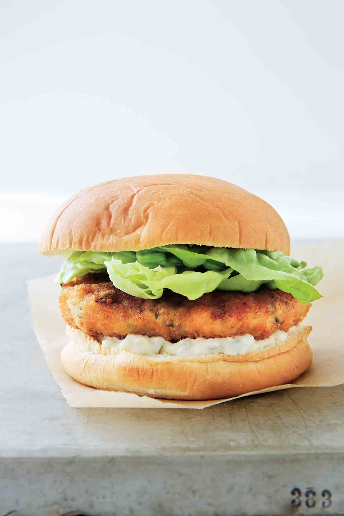 A shrimp burger with tartar sauce, topped with lettuce in a white bun on a piece of parchment paper.