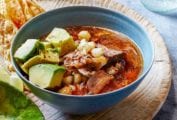 A bowl of pork, avocado, and hominy in a red broth on a cutting board with tortilla chips