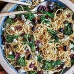 A plate of spaghetti with olives, broccoli, and feta with two serving spoons nestled in the pasta.
