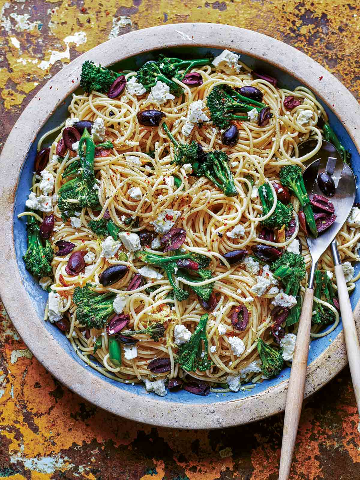 A plate of spaghetti with olives, broccoli, and feta with two serving spoons nestled in the pasta.