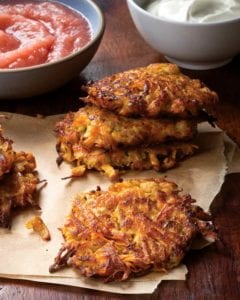 Several sweet potato and apple latkes on a paper towel with a bowl of applesauce in the background.