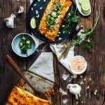 A casserole dish of sweet potato enchiladas with a place with two enchiladas, lime, jalapenos, and sauce