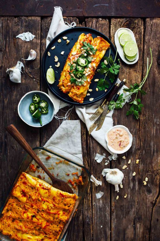 A casserole dish of sweet potato enchiladas with a place with two enchiladas, lime, jalapenos, and sauce.