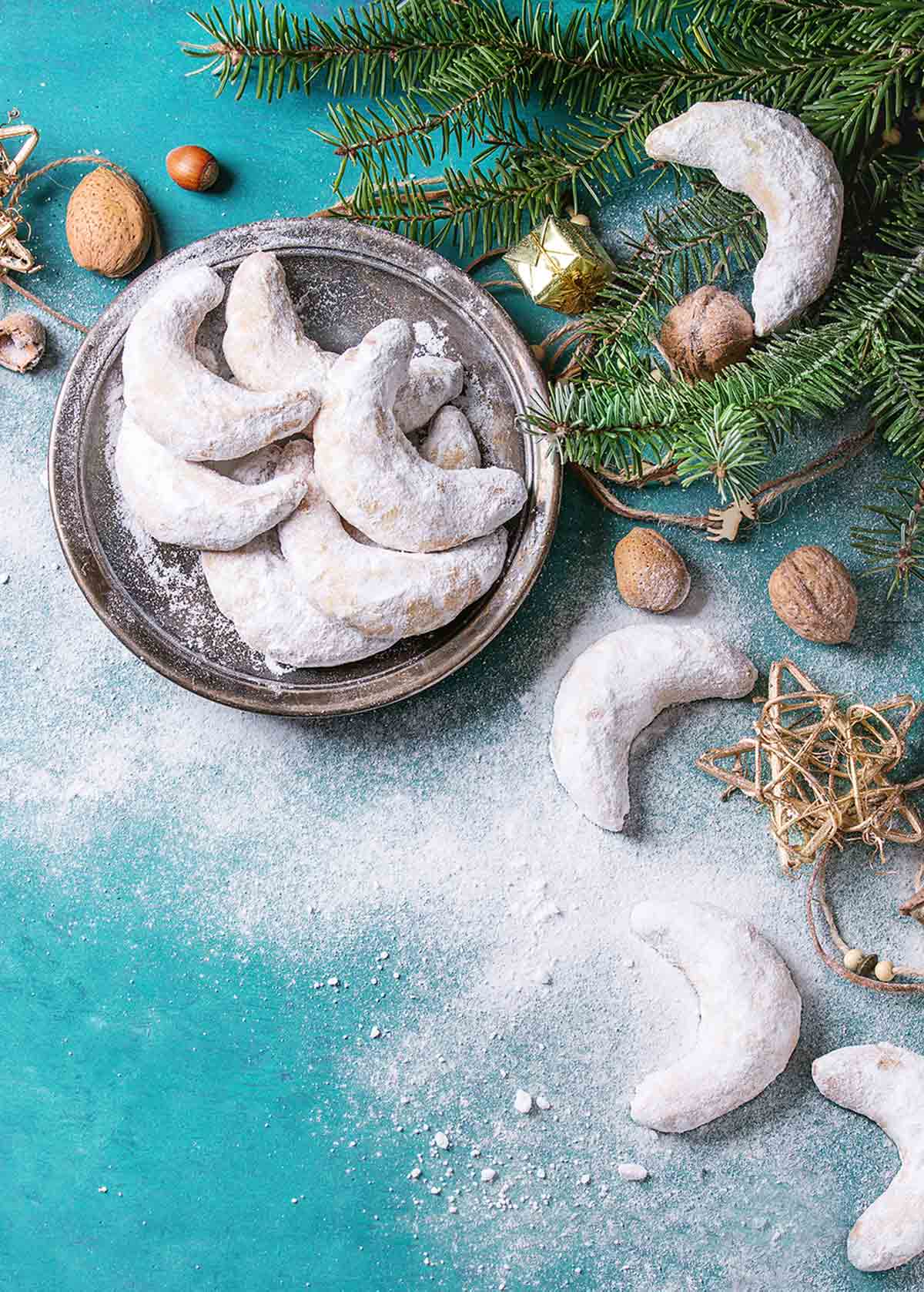 A pile of crescent-shaped cookies on a platter with greenery and Christmas ornaments on the side