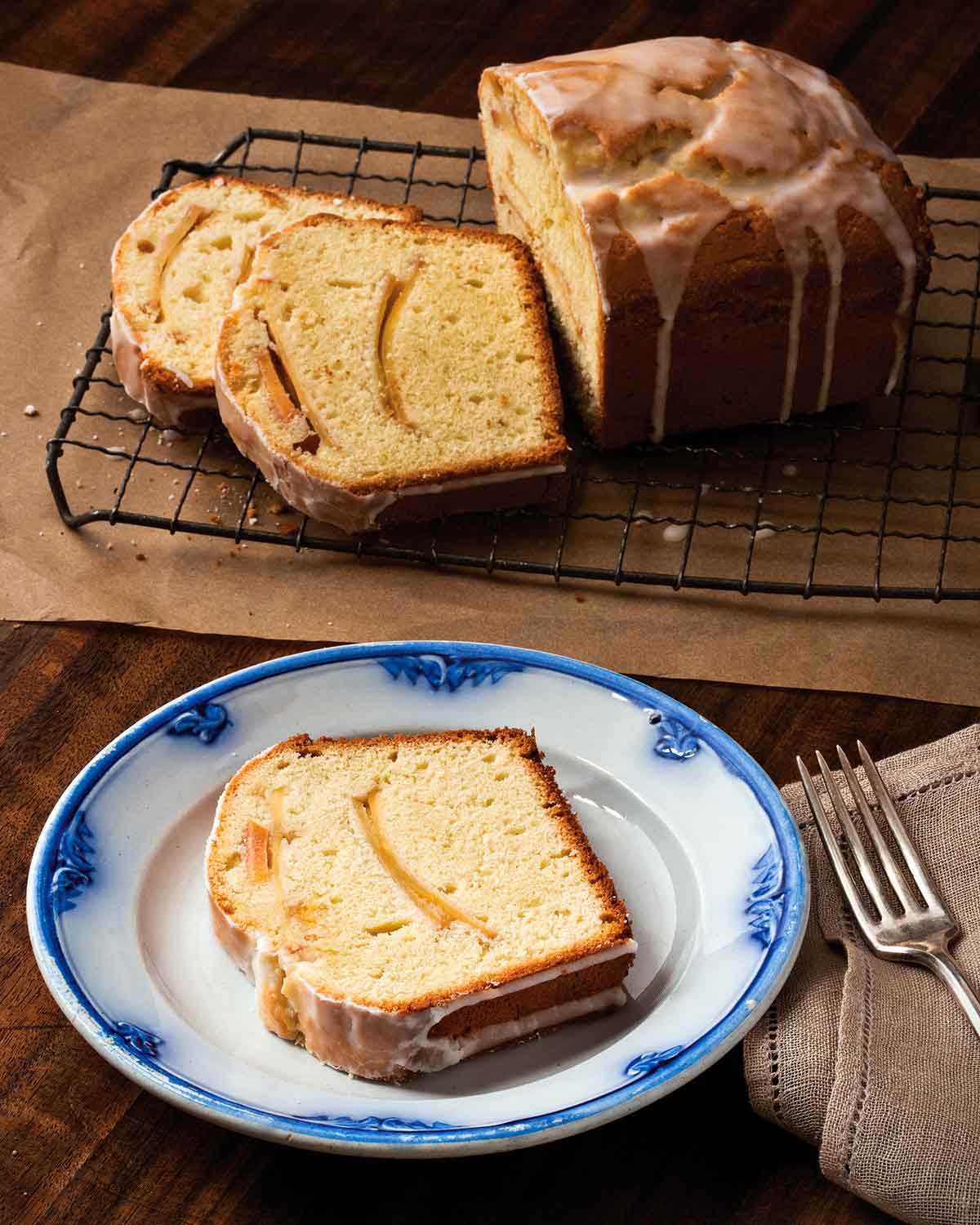 An apple tea cake on a wire rack with three slices cut from it and one on a blue and white plate.