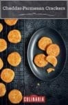 Cheddar-Parmesan crackers on a black wire rack with a few on a black oval plate beside the rack.