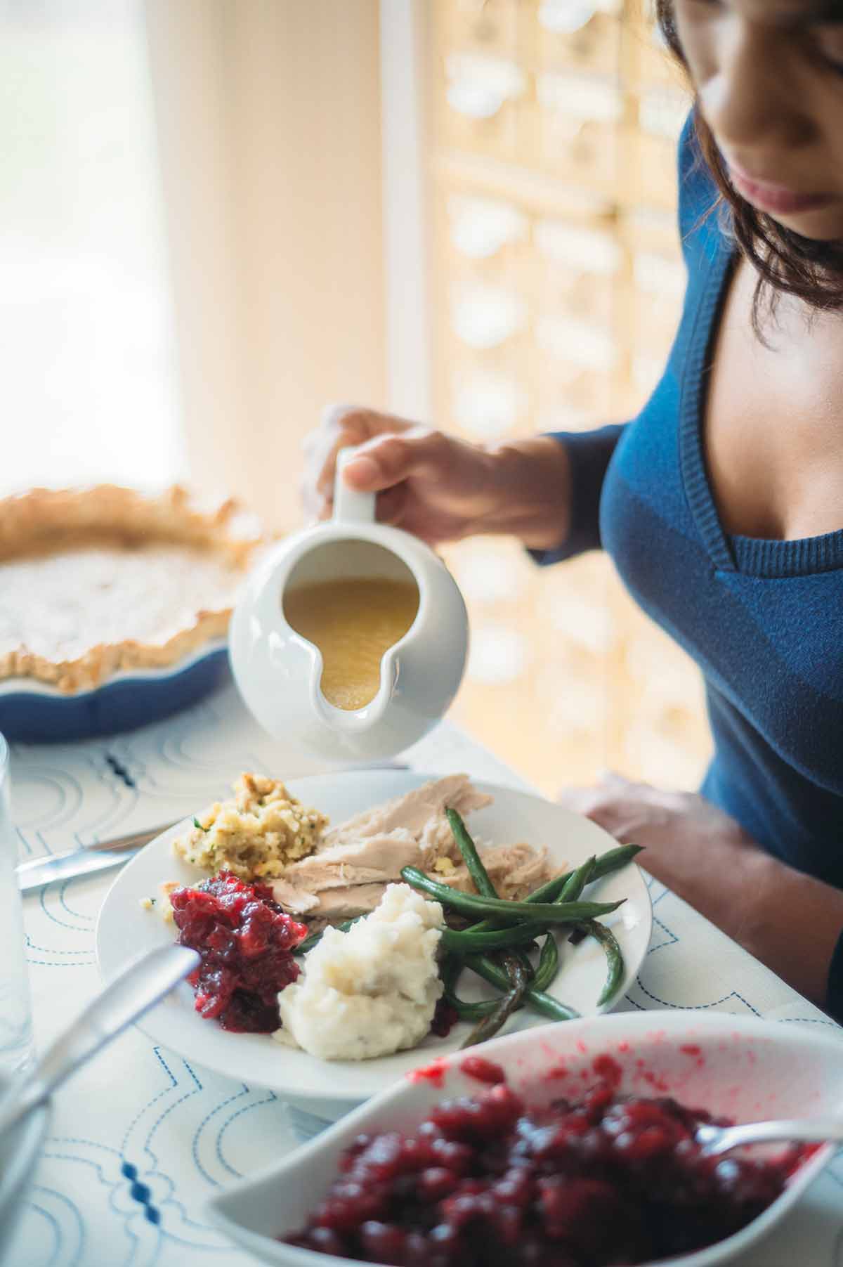 A person holding a gravy boat over a full plate of Thanksgiving dinnner.