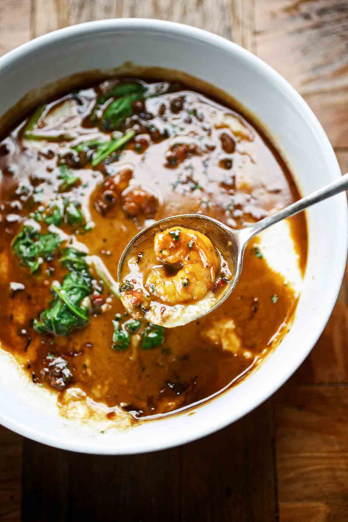 Creole shrimp and grits in a white bowl, with a spoon above the bowl with a shrimp in it.