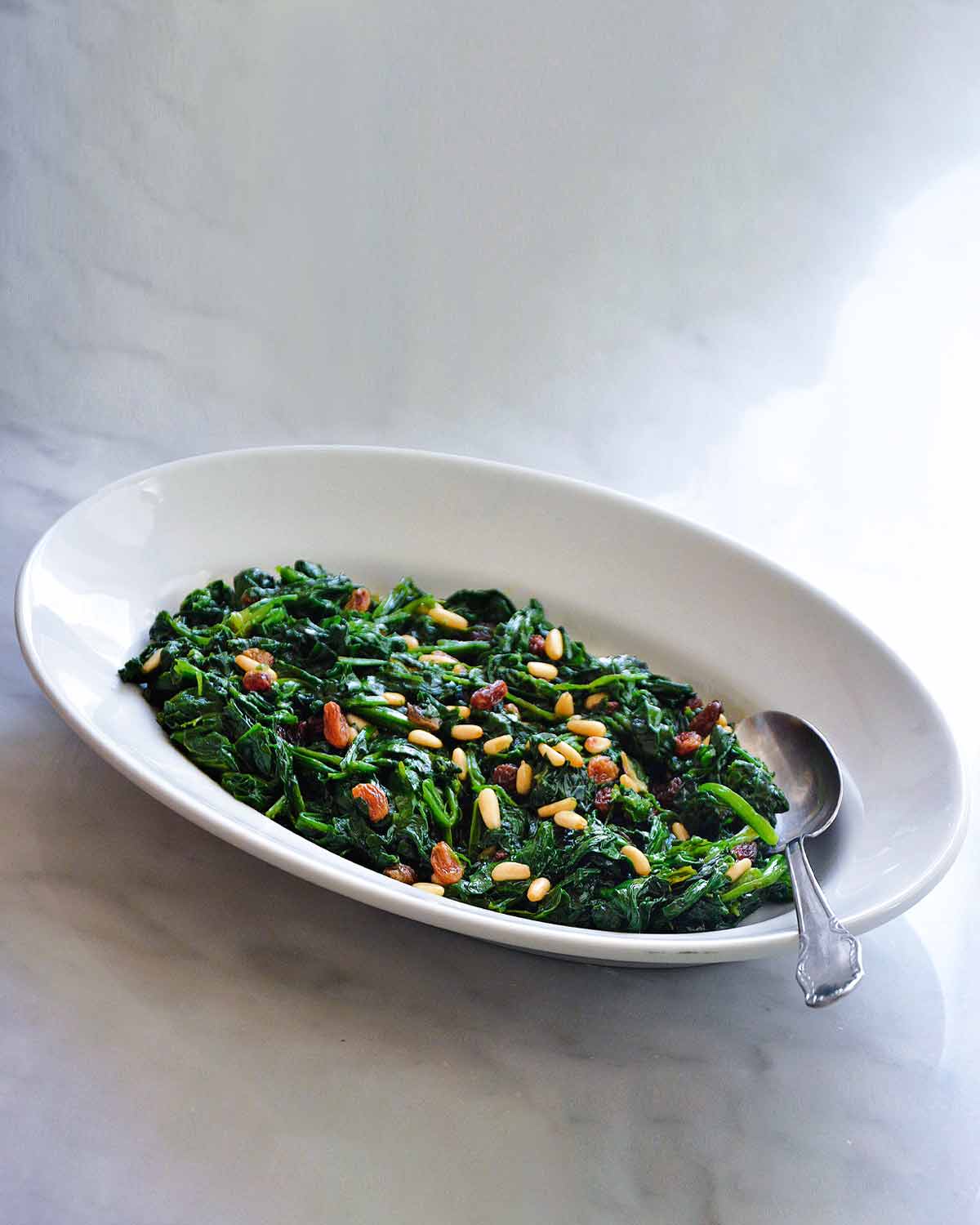 A white oval dish filled with cooked spinach with raisins and pine nuts sprinkled over the top and a spoon resting in the dish.