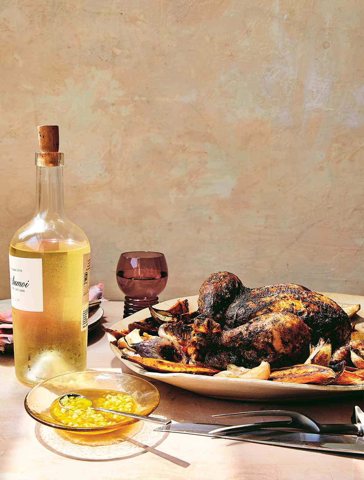 A serving platter topped with baharat roast chicken with sweet potatoes and a bowl of preserved lemon oil on the side along with a bottle of wine and a wine goblet.