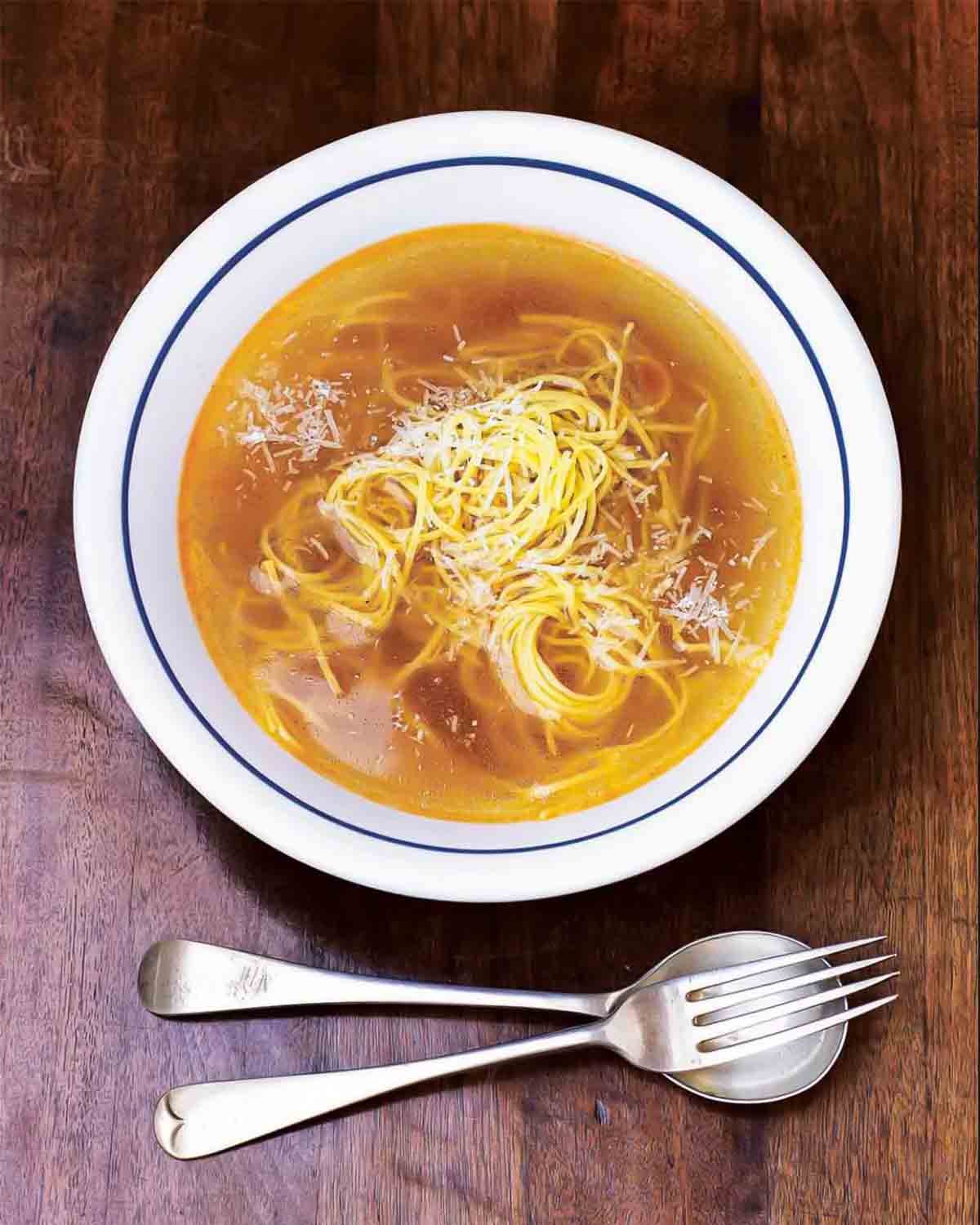 A white bowl filled with brodo di pollo on a wooden surface with a fork and spoon beside the bowl.