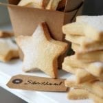 A stack of star-shaped cutout classic shortbread cookies, lightly dusted with sugar, and a brown takeout container and a gift tag.