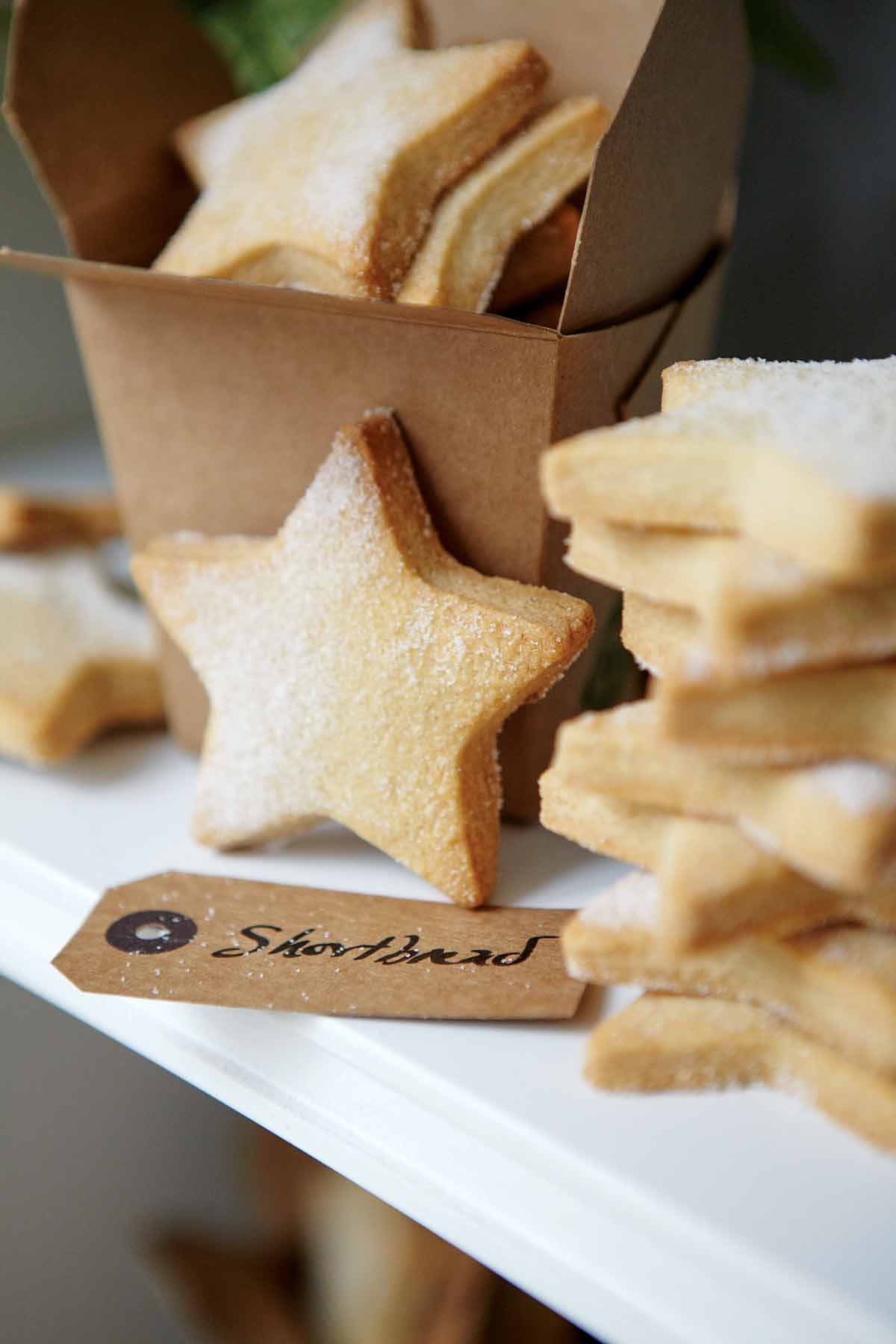 A stack of star-shaped cutout classic shortbread cookies, lightly dusted with sugar, and a brown takeout container and a gift tag.