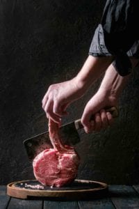 A person holding a cleaver and demonstrating how to french trim a roast.