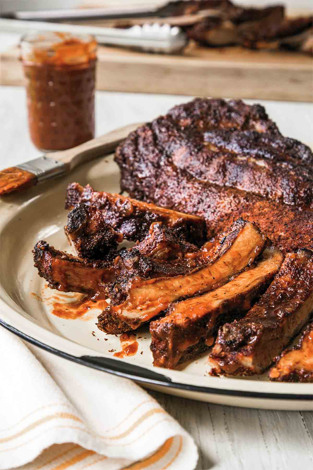 A rack of oven baked barbecue ribs on a white platter with a jar of sauce and a brush next to them.