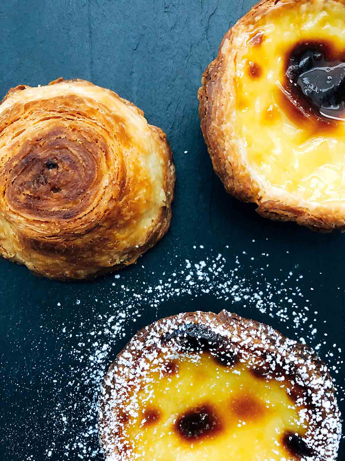 Three pasteis de nata on a slate background sprinkled with powder sugar.