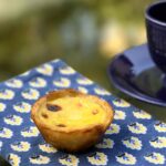 One pasteis de nata on a flowered napkin with a coffee cup nearby.