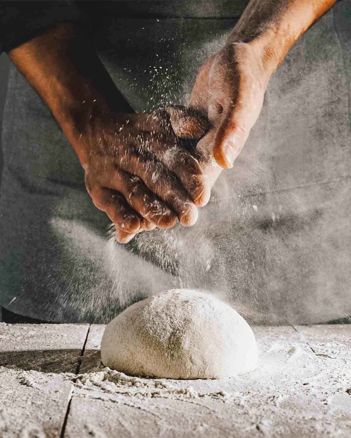 A person dusting flour over a ball of quick pizza dough on a wooden table.