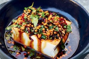 A piece of silken tofu with pine nuts and pickled chiles topped with cilantro in a blue ceramic bowl.