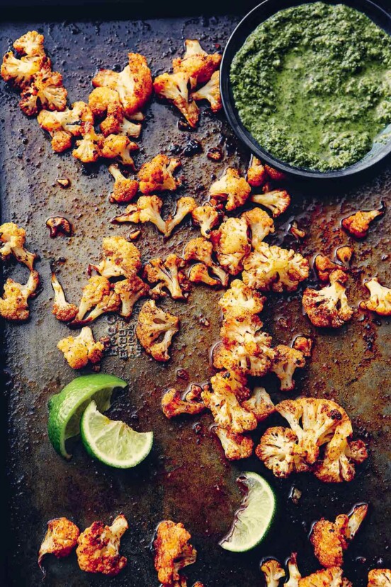 Florets of spicy roasted cauliflower on a baking sheet with squeezed lime wedges and a bowl of green sauce beside them.