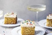 Squares of vanilla sheet cake with Swiss buttercream frosting and sprinkles on white plates with a coupe of Champagne in the background.