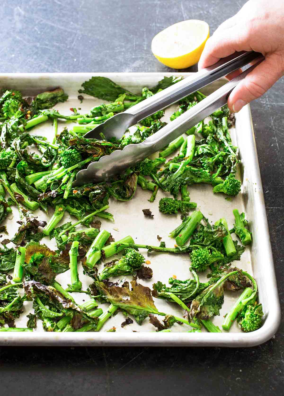 Pieces of broiled broccoli rabe being turned over on a sheet pan by a person using tongs.