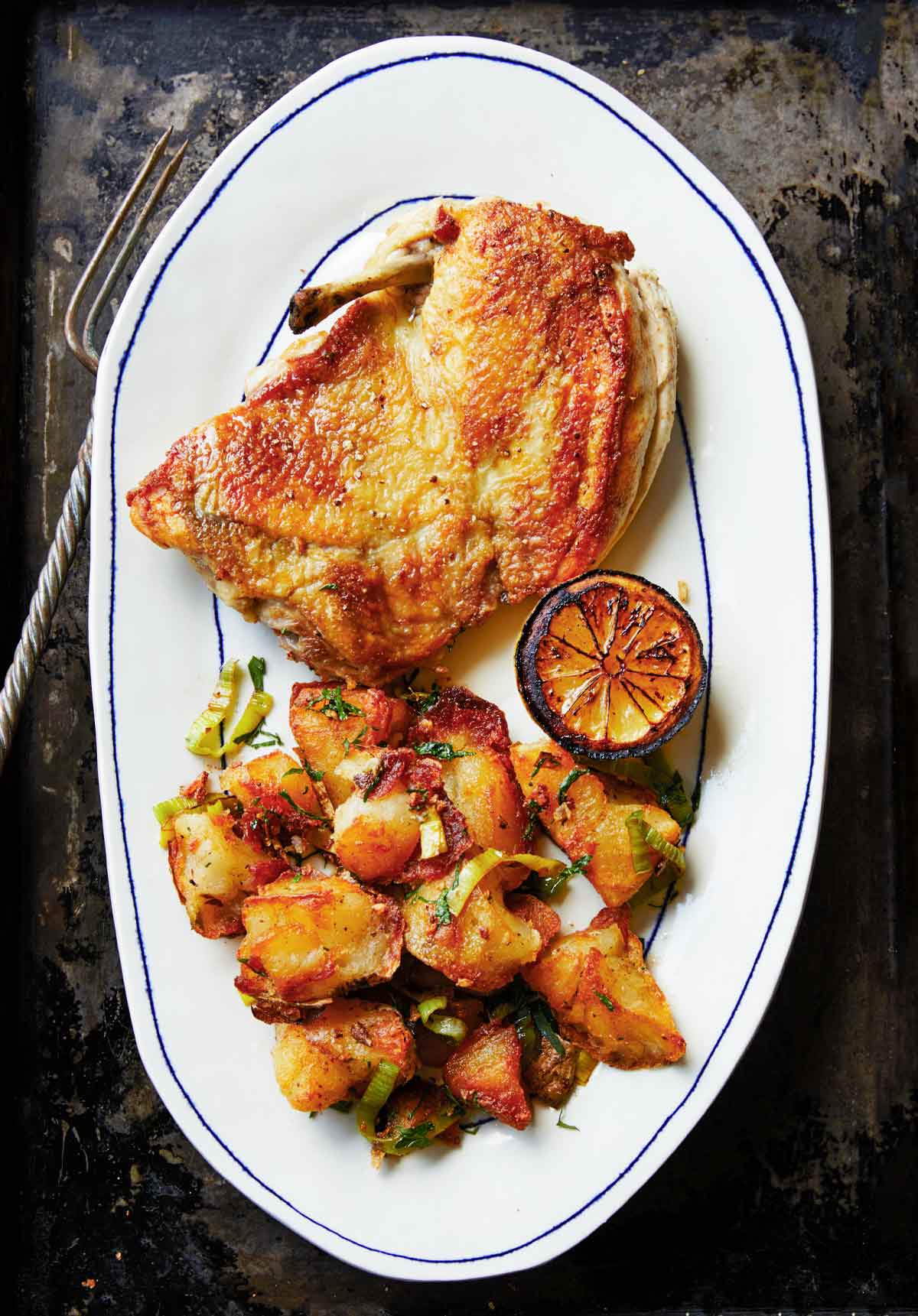 A piece of crisp roast chicken in a skillet on a plate with crispy potatoes and charred lemon.