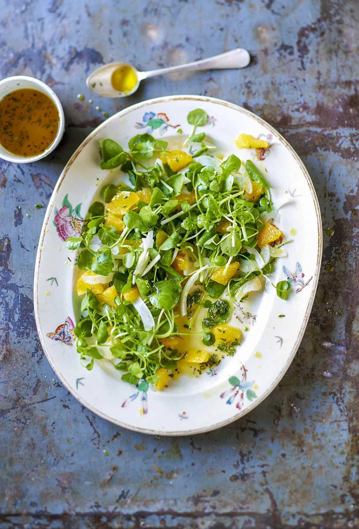 A fennel, orange, and watercress salad on a decorative platter with a cup of dressing and a spoon on the side.