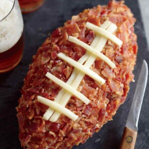 A football cheese ball on a serving board with two glasses of beer and a knife beside it.
