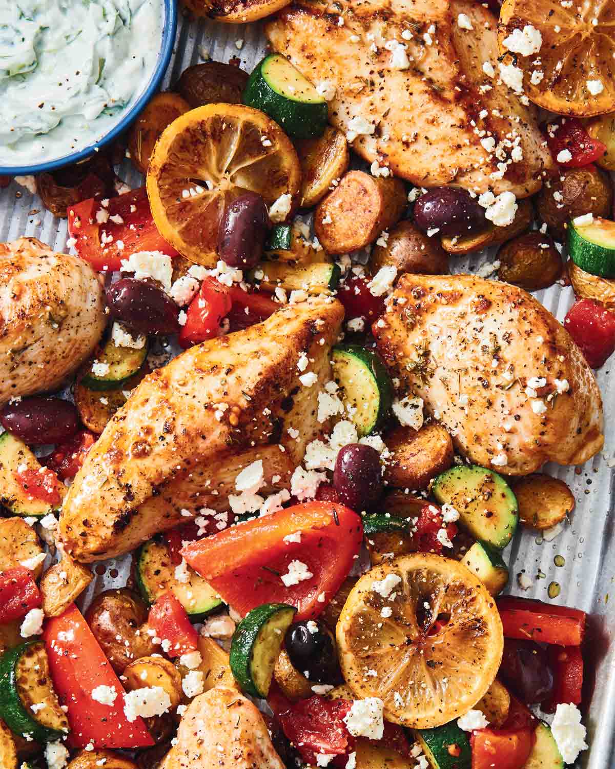 Greek-style sheet pan chicken, olives, zucchini, and lemon slices on a sheet pan with a bowl of tzatziki nearby.