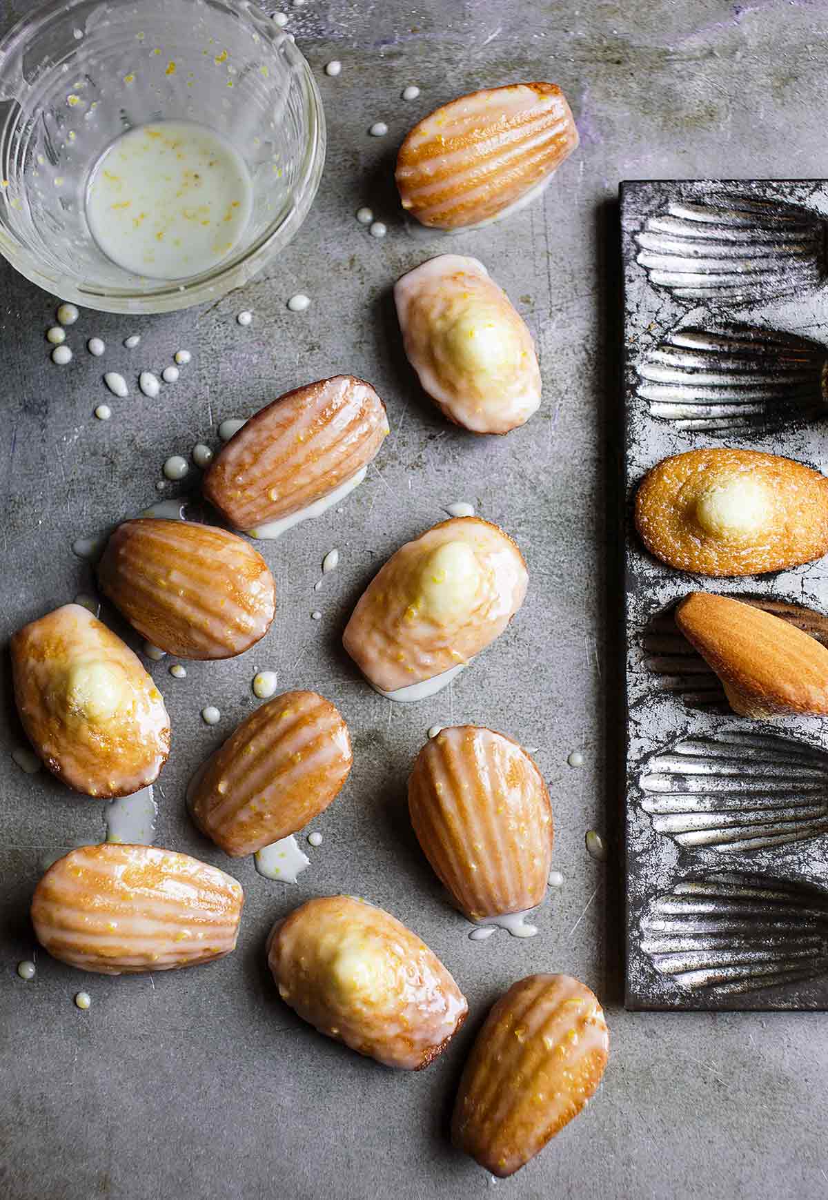 Several lemon madeleines next to a madeleine pan and a bowl of lemon glaze nearby.