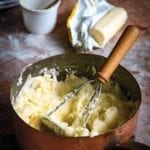 A copper pot filled with mashed potatoes with crème fraîche and a masher resting inside.