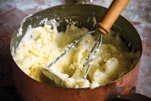 A copper pot filled with mashed potatoes with crème fraîche and a masher resting inside.