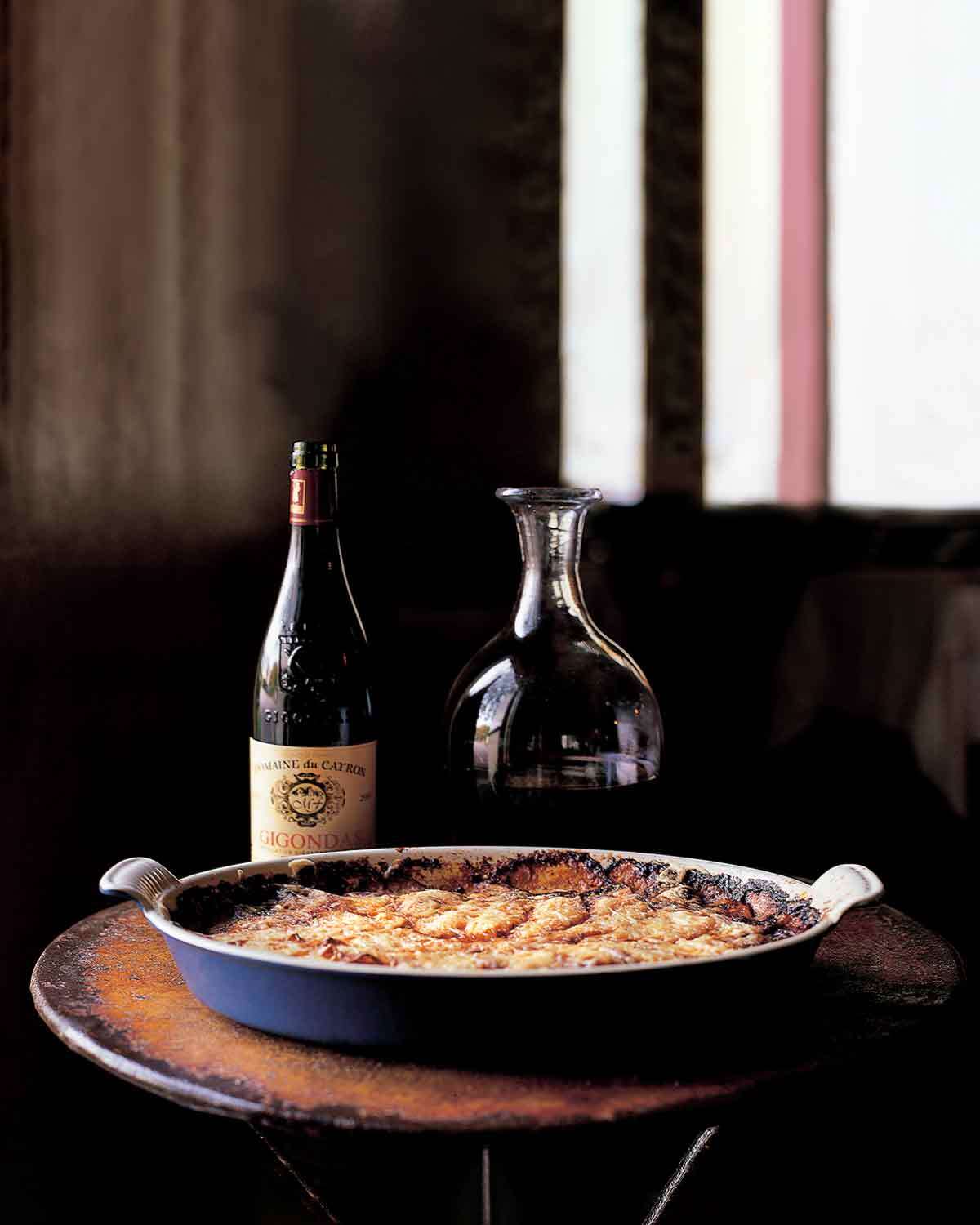 An oval dish of old-fashioned potato gratin, with a bottle and decanter of wine behind it.