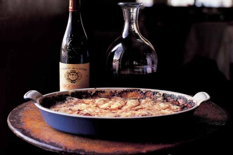 An oval dish of old-fashioned potato gratin, with a bottle and decanter of wine behind it.