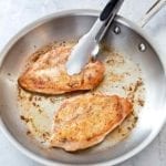 Two pan seared chicken breasts in a stainless steel skillet with a pair of tongs resting on one of the chicken breasts.