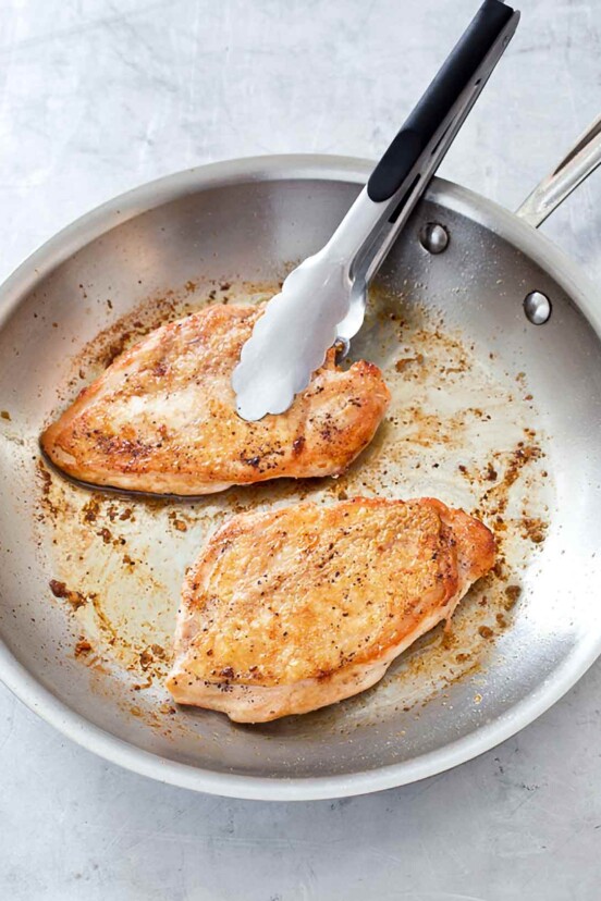 Two pan seared chicken breasts in a stainless steel skillet with a pair of tongs resting on one of the chicken breasts.