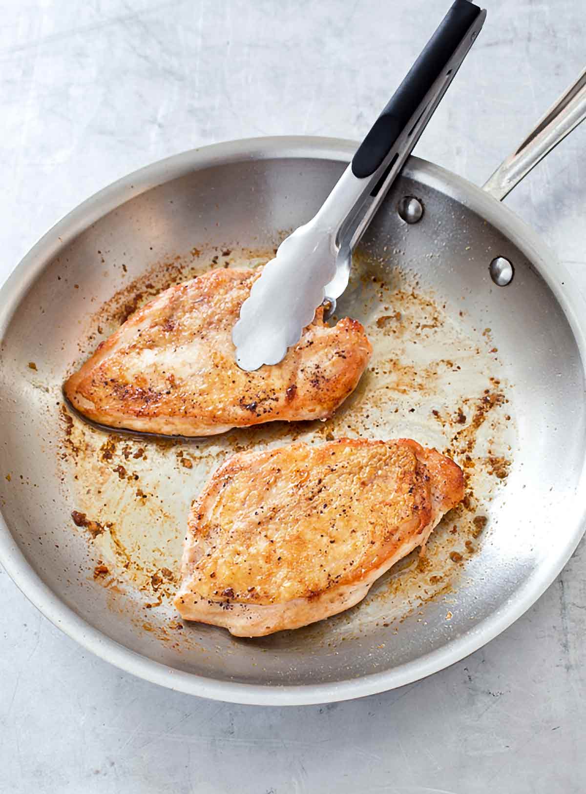 Two pan seared chicken breasts in a stainless steel skillet with a pair of tongs resting on one of the chicken breasts.