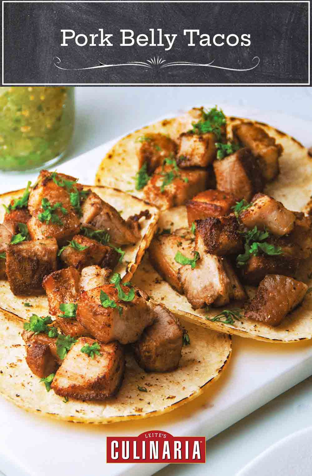 Four pork belly tacos garnished with cilantro and a jar of salsa in the background.