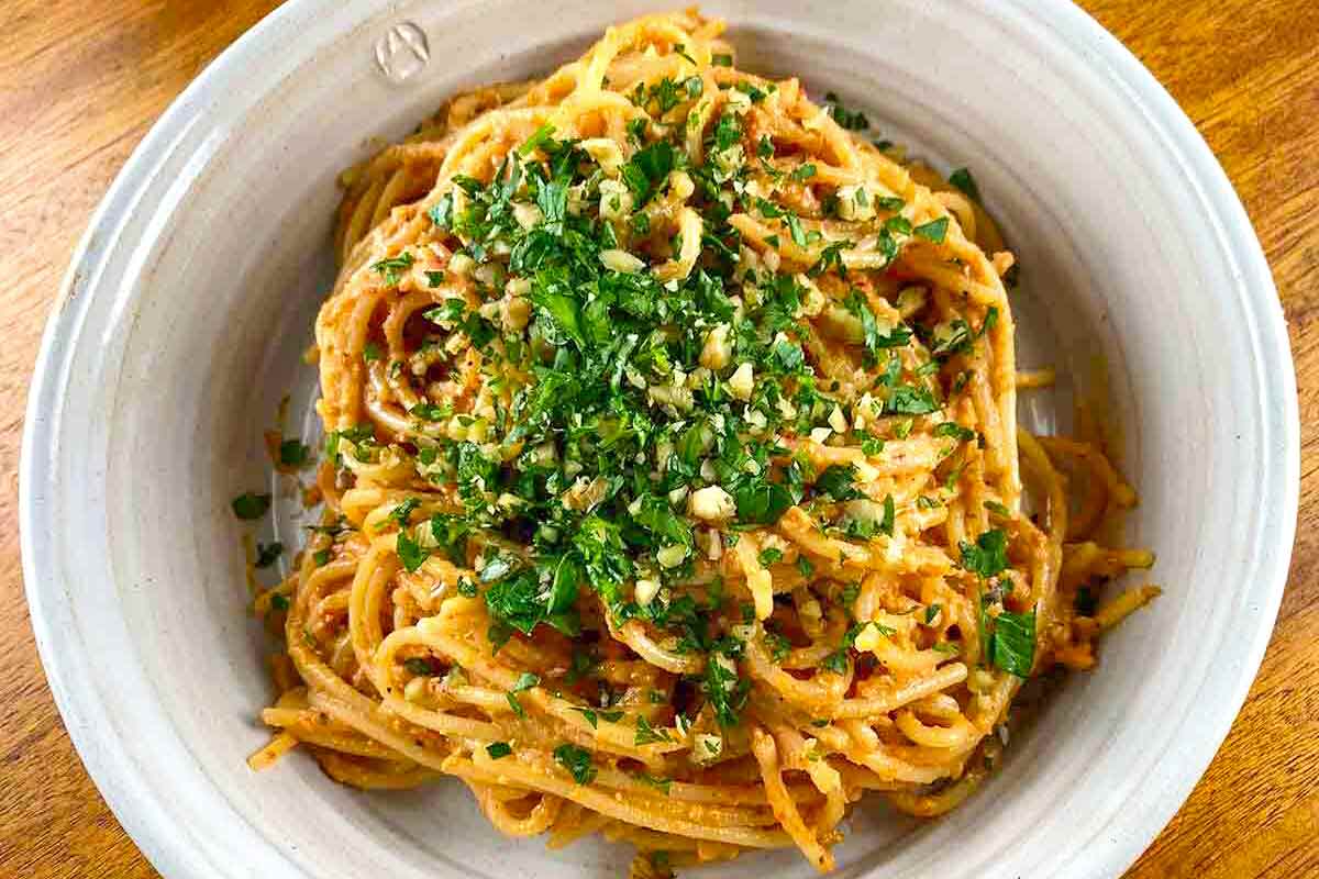 A bowl of spaghetti with muhammara sauce, topped with parsley and walnuts.