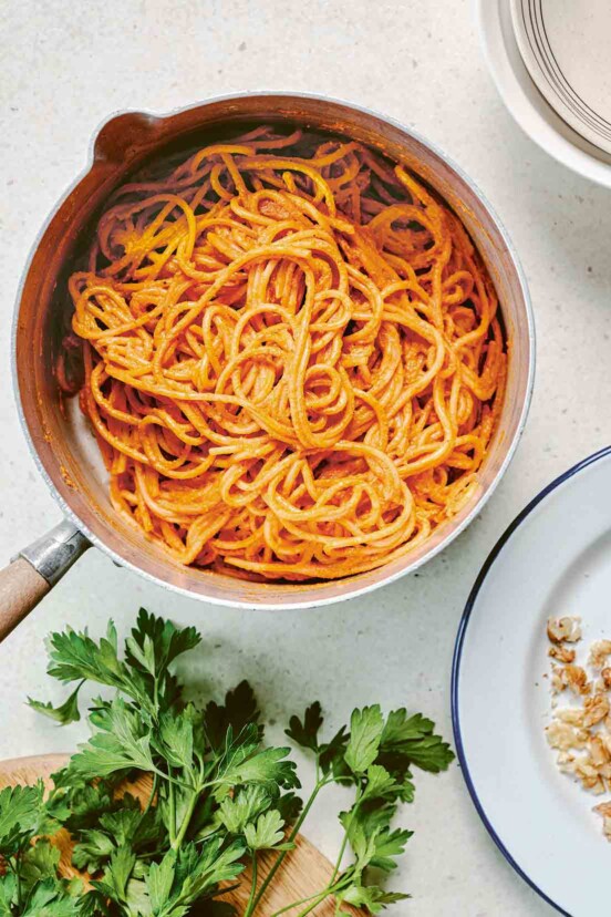 A pot of spaghetti with muhammara sauce next to a plate and a cutting board with parsley on it.
