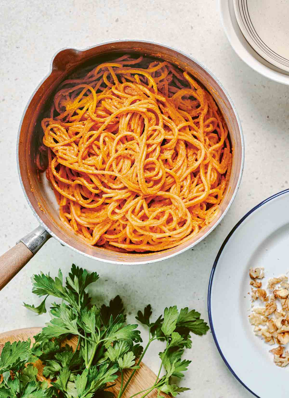 A pot of spaghetti with muhammara sauce next to a plate and a cutting board with parsley on it.