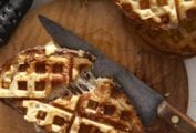 Two waffle iron grilled cheese sandwiches on a wooden cutting board with a knife and a waffle iron nearby.