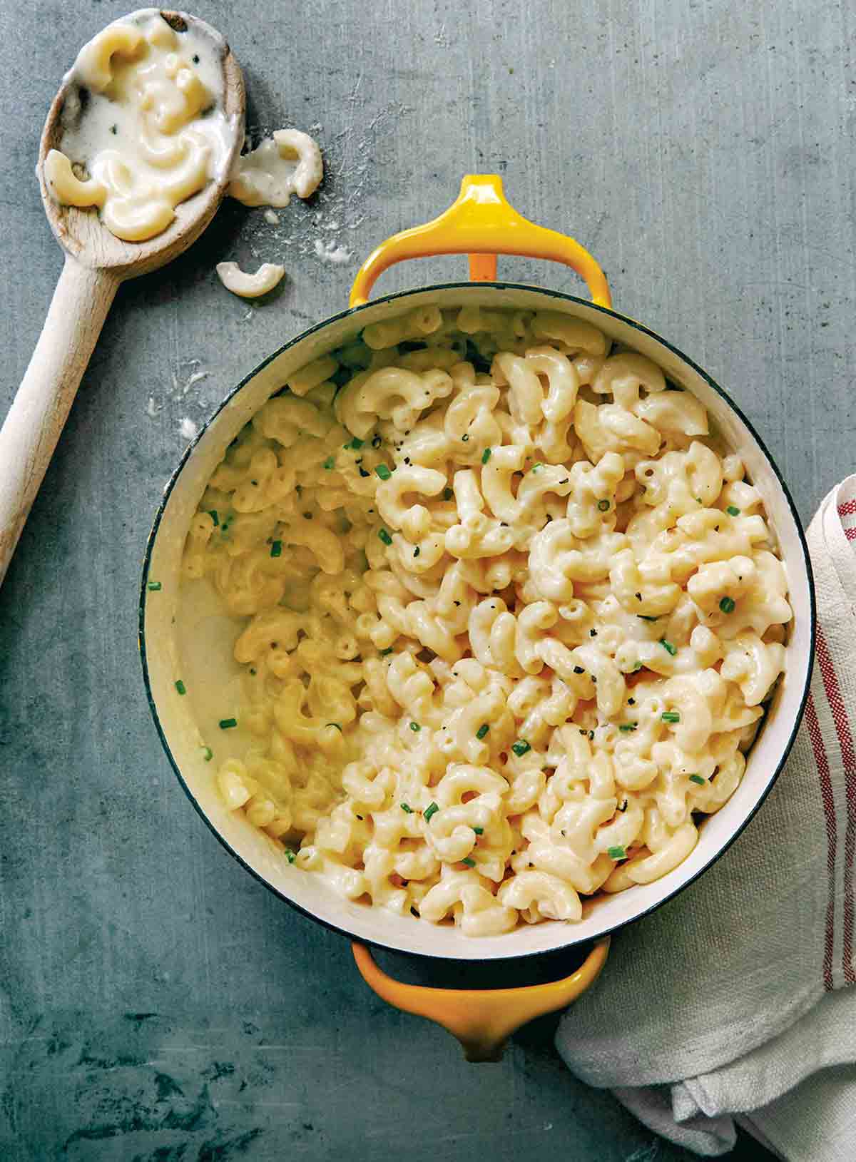 A creamy pot of three-ingredient macaroni and cheese in a yellow Dutch oven, garnished with chives. A wooden spoon rests nearby with a bit of cheesy pasta on it.