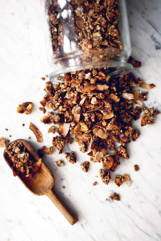 Banana bread granola tipped out of a glass jar onto a marble surface with a wooden scoop beside it.