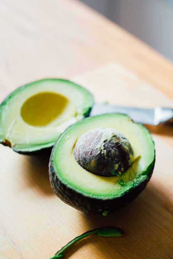 A halved avocado on a wooden board with a knife beside it for the writing 'I Tried Blanching Avocados. Here's What Happened.'