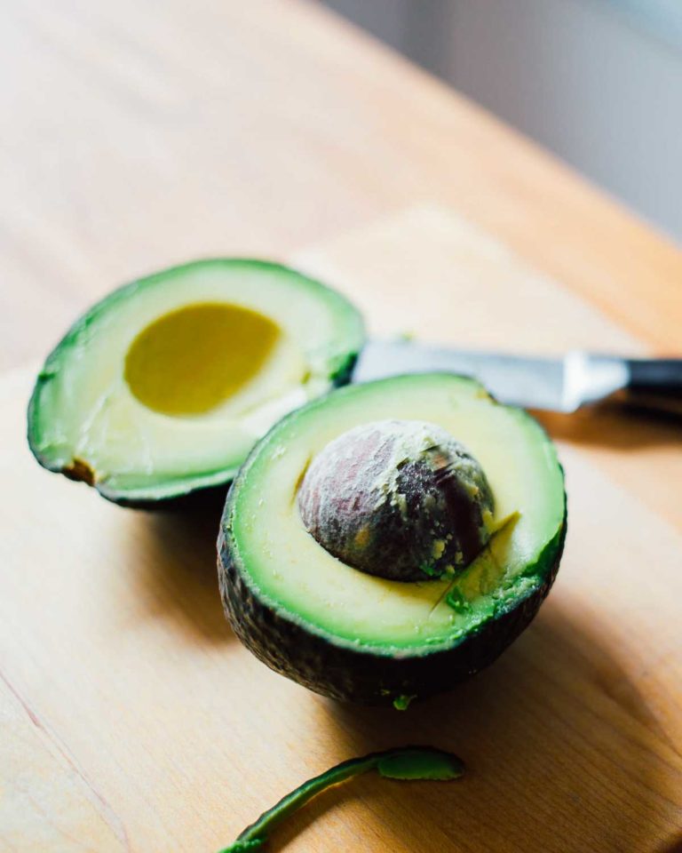 A halved avocado on a wooden board with a knife beside it for the writing 'I Tried Blanching Avocados. Here's What Happened.'