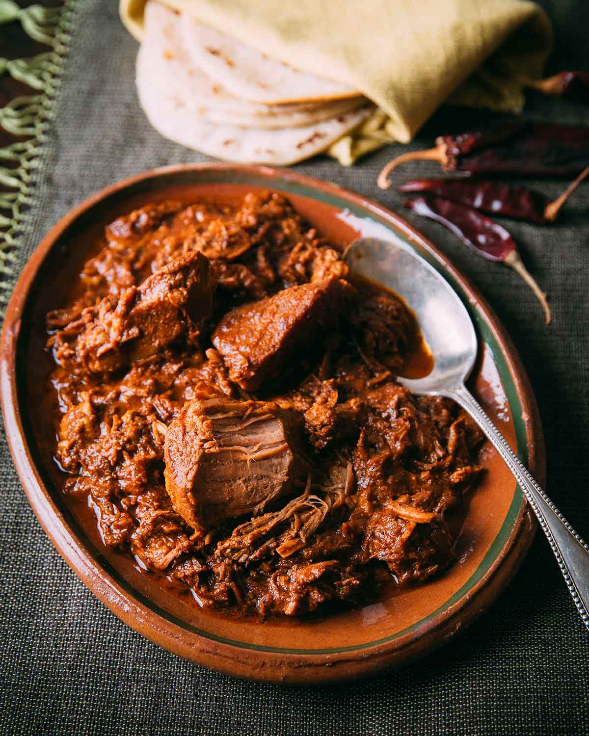 An oval bowl filled with braised pork in red chile sauce with a spoon resting on the side and a stack of tortillas and dried peppers on the side.